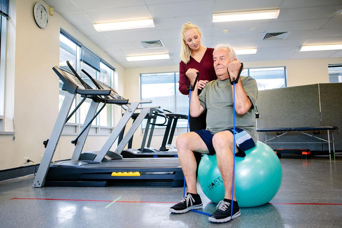 Man sitting on physio ball with support from physiotherapist.